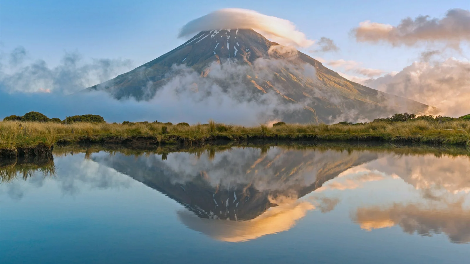 一座云雾缭绕的山峰倒映在平静的湖面上，前景是草地与水岸。