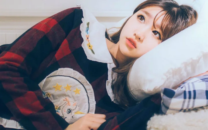 一名女性穿着格纹上衣侧躺在床上，头靠着白色枕头看向镜头。