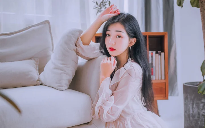 一名成年女性穿着浅色上衣，坐在沙发旁的室内空间中。