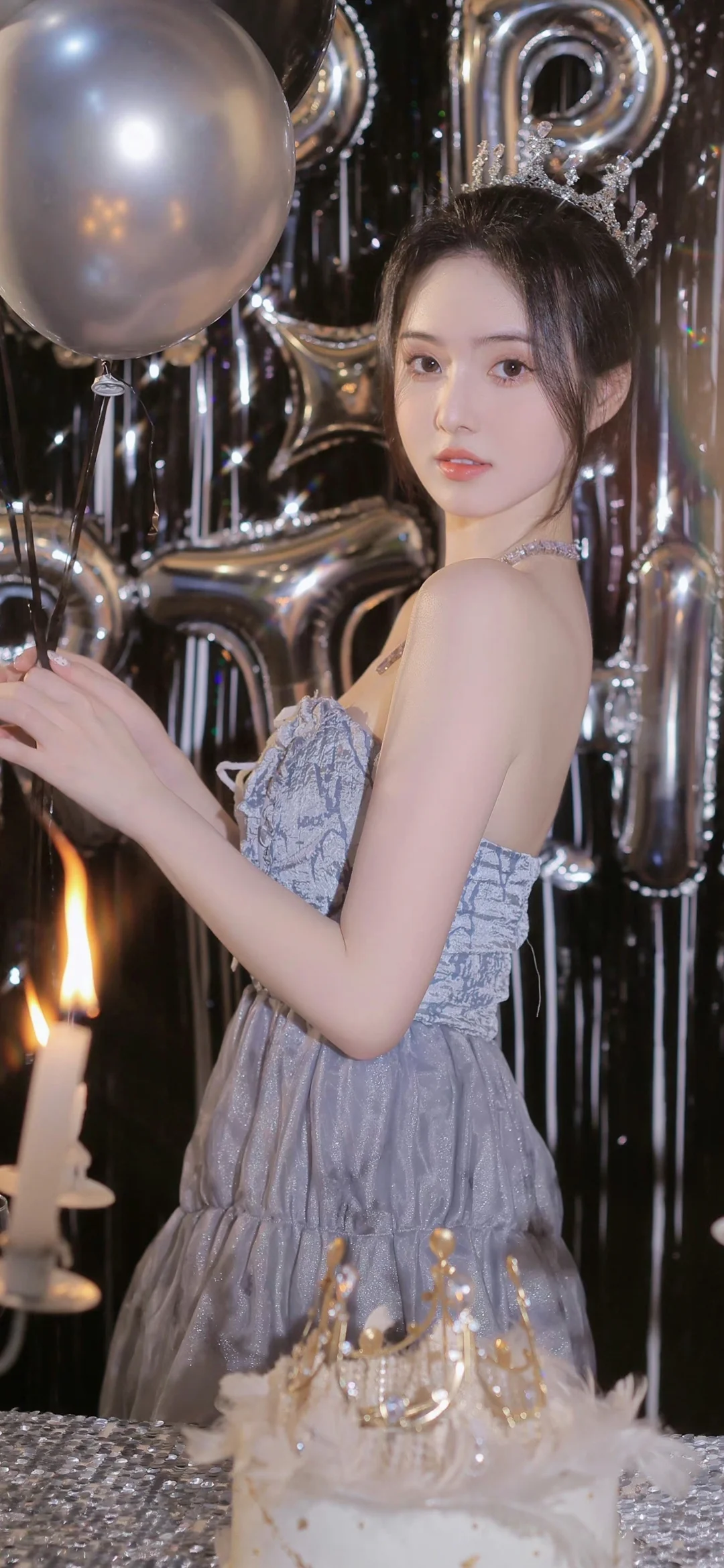 一位身穿银灰色礼服的女性站在银色气球和生日蛋糕前。