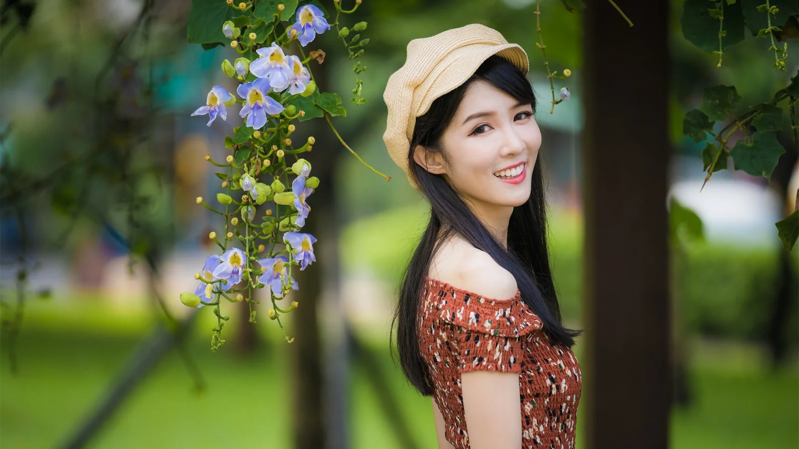 一位戴着米色帽子的女子站在花园中紫色花朵旁微笑。