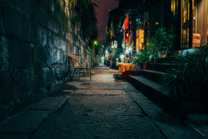夜晚的城市小巷中，石墙、店铺灯光、植物和长椅沿着狭窄街道延伸。