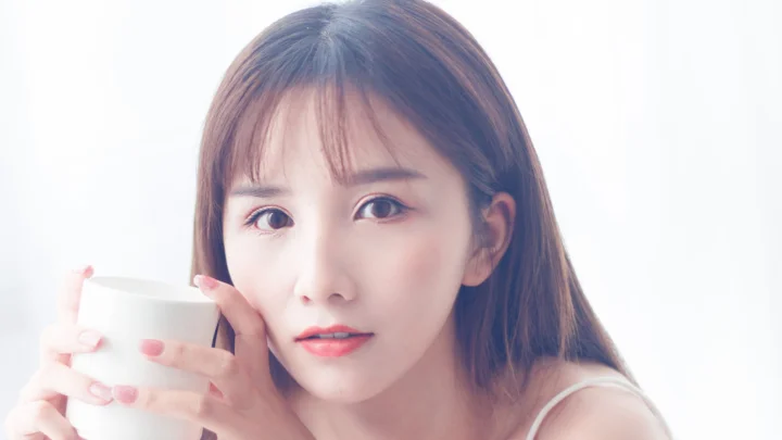 밝고 단순한 배경에서 흰색 컵을 들고 카메라를 바라보는 여성의 클로즈업 인물 사진입니다.