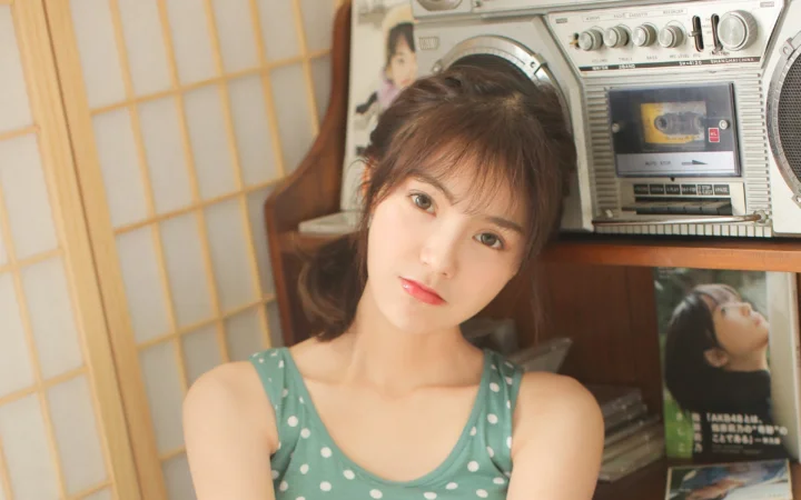 초록색 물방울무늬 상의를 입은 성인 여성이 부드러운 실내 조명 아래 빈티지 라디오 옆에 앉아 있습니다.