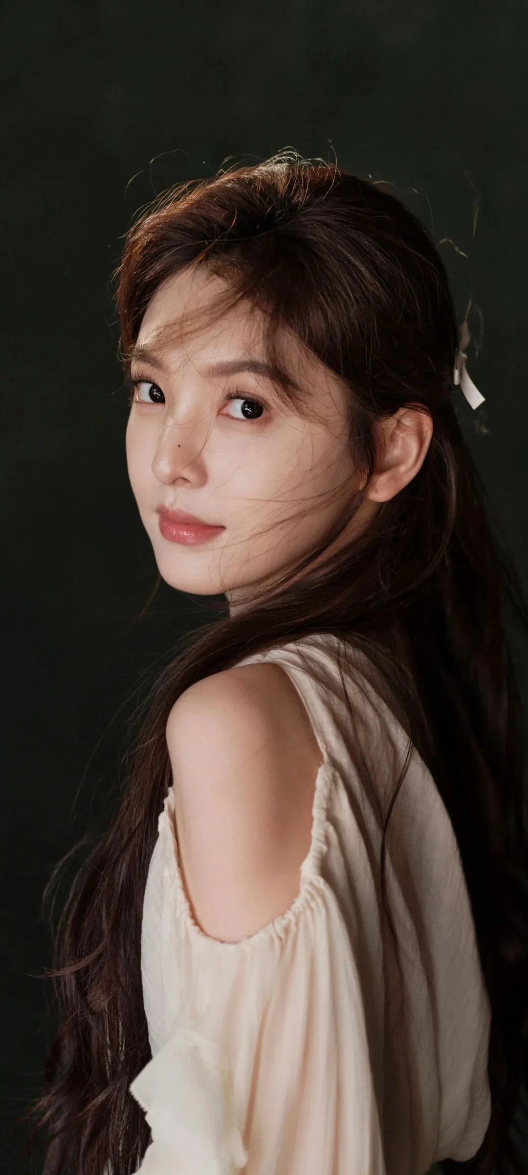 긴 머리의 여성이 어두운 배경을 뒤로하고 크림색 오프숄더 상의를 입은 채 돌아보며 카메라를 바라보고 있다.