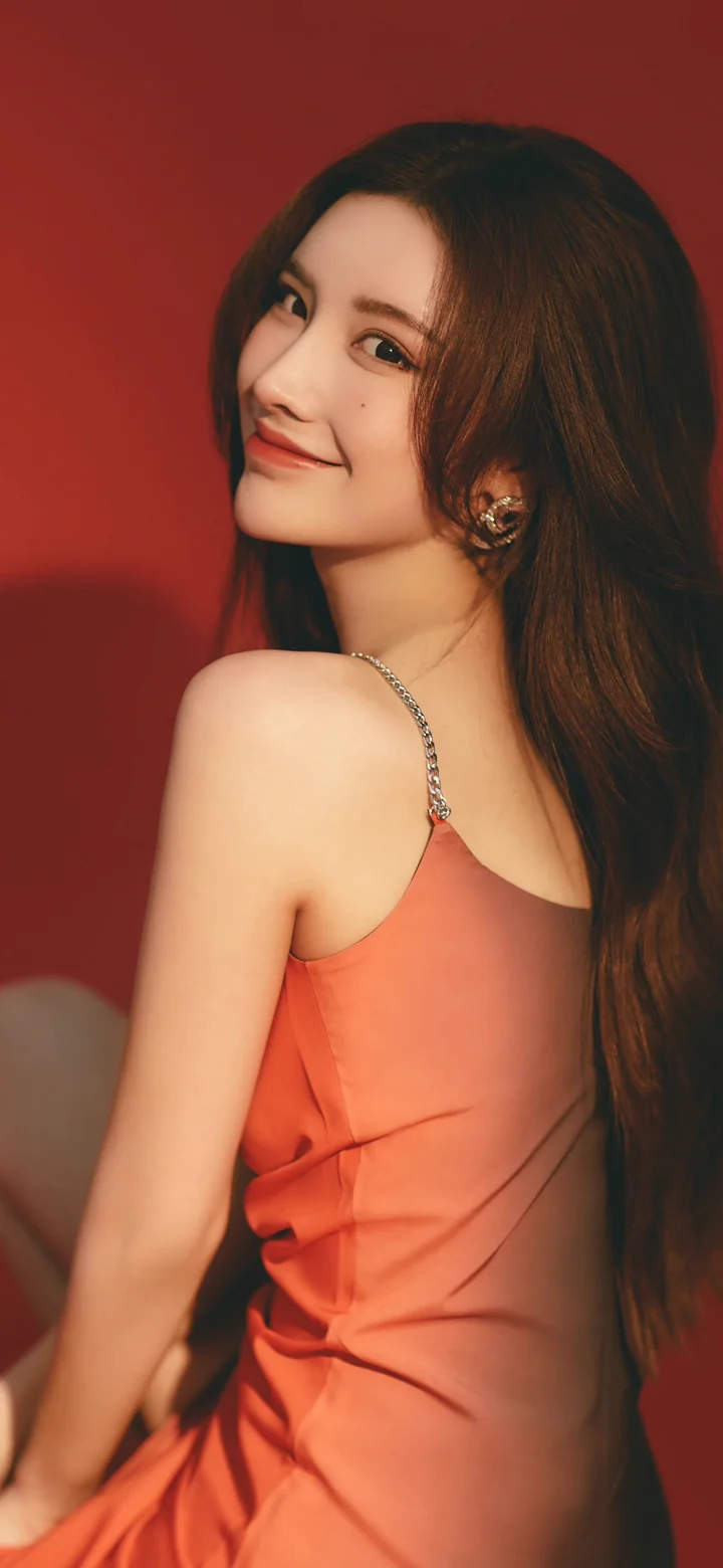 붉은 배경 앞에서 오렌지색 끈 드레스를 입은 긴 머리 여성이 뒤돌아보며 미소 짓고 있다.