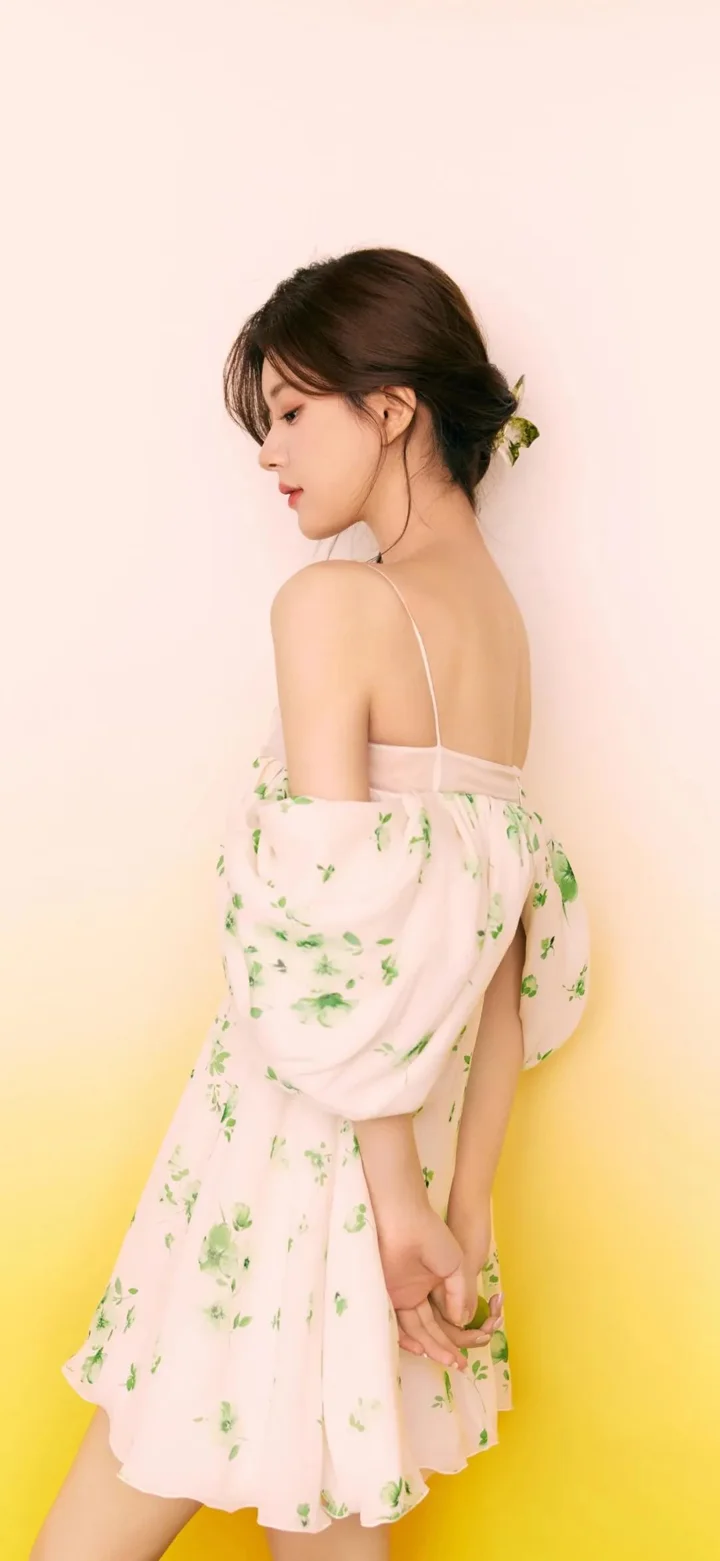 一名成年女性身穿粉色碎花连衣裙，在浅粉色背景前侧身回望。