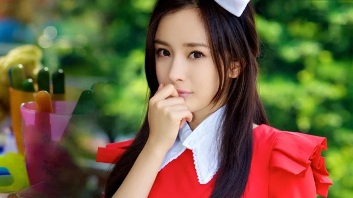 一名长发女性身穿红色服装，在绿色户外背景前看向镜头。