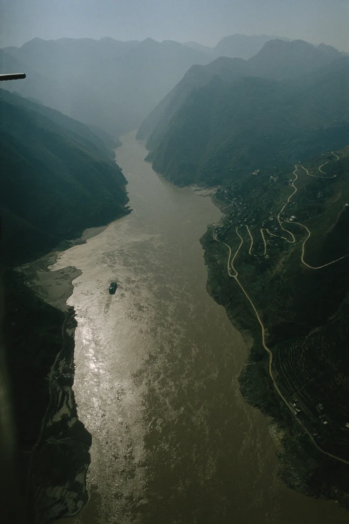航拍视角展现一条河流穿过群山峡谷，山坡上可见蜿蜒道路。