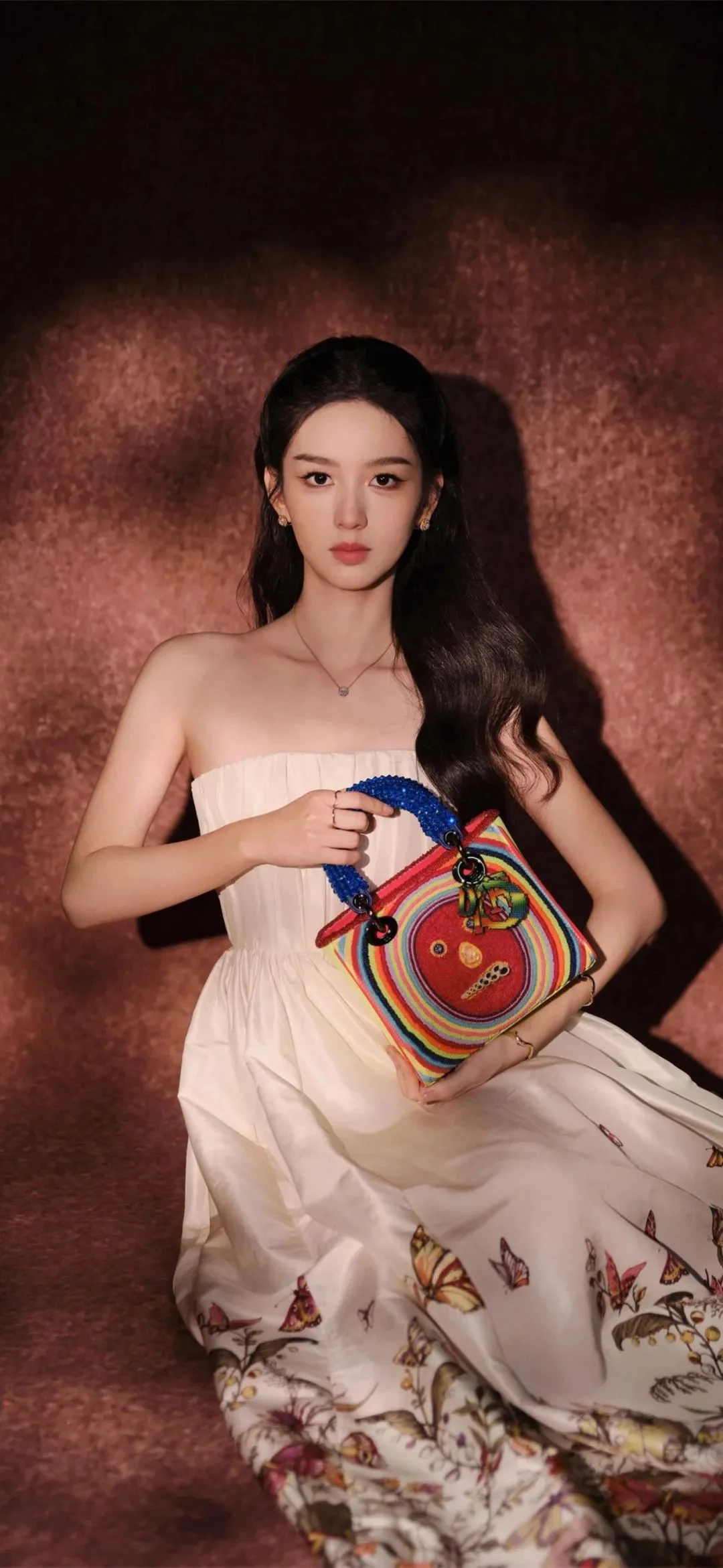 一名成年女性身穿白色印花长裙，手提彩色手袋，在棕色背景前摆姿势。