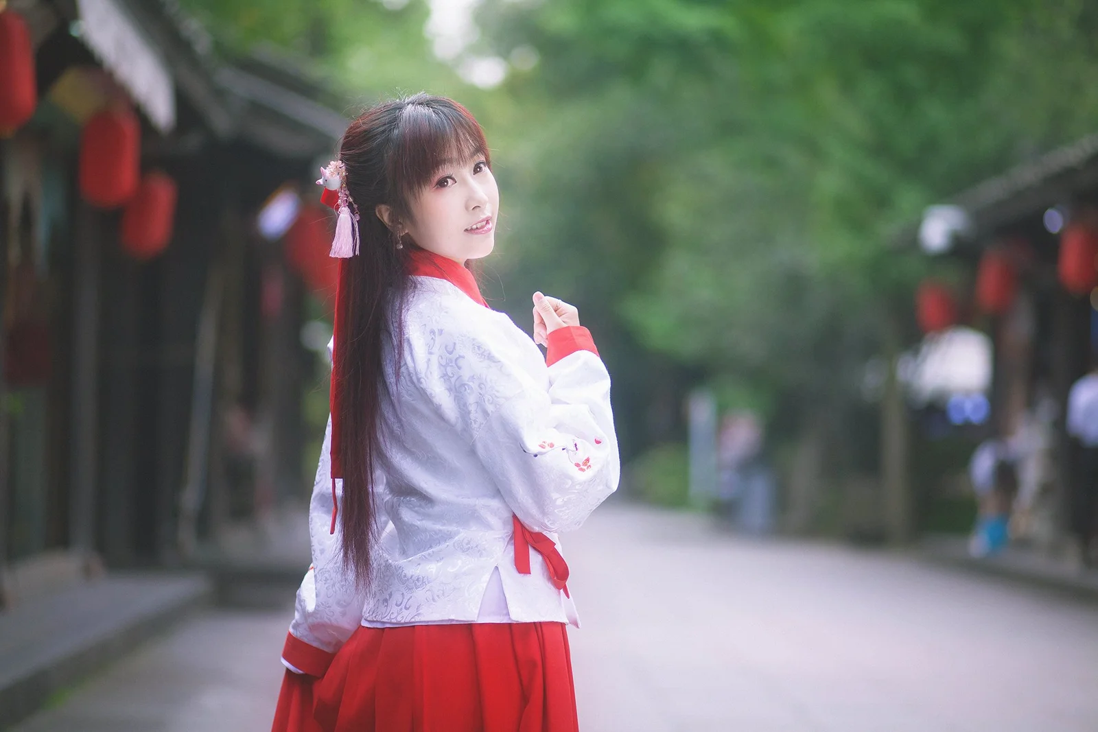 一名身穿红白传统服饰的女子站在林荫街道上回望镜头。