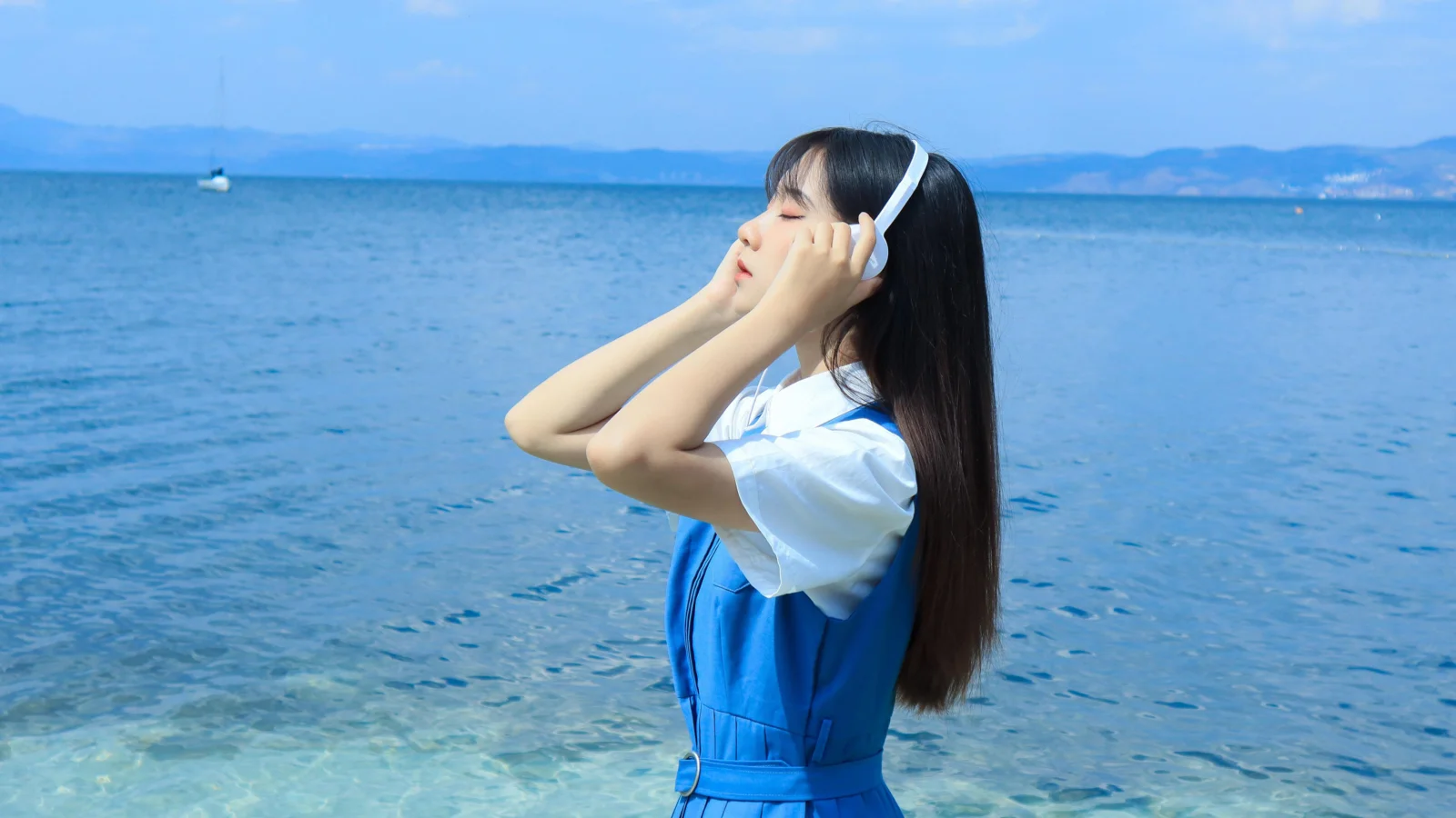 一名戴着耳机的女子站在清澈的海水旁，远处是蓝色海面与山脉。