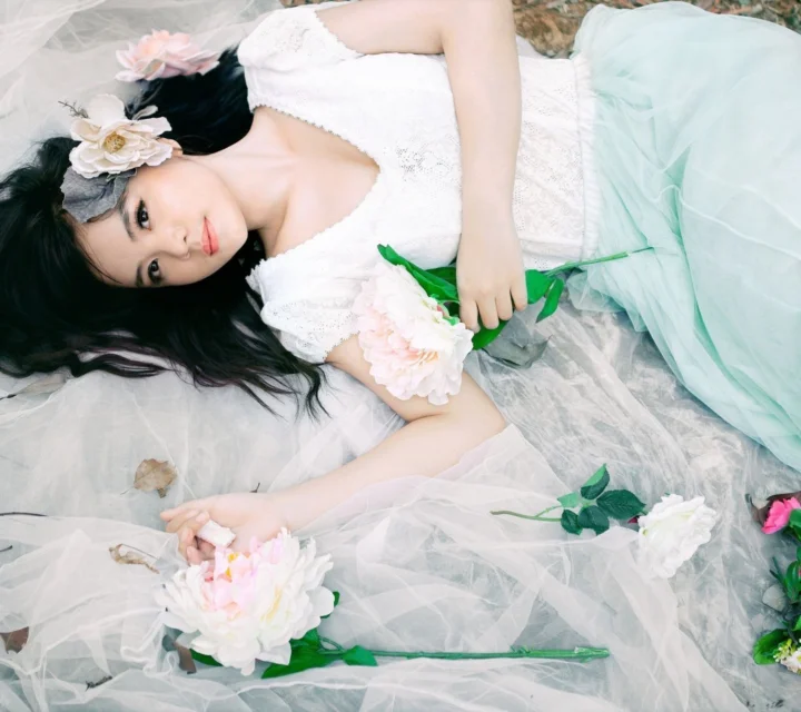 흰 드레스를 입은 여성이 얇은 천과 꽃 사이에 누워 꽃다발을 들고 있다.