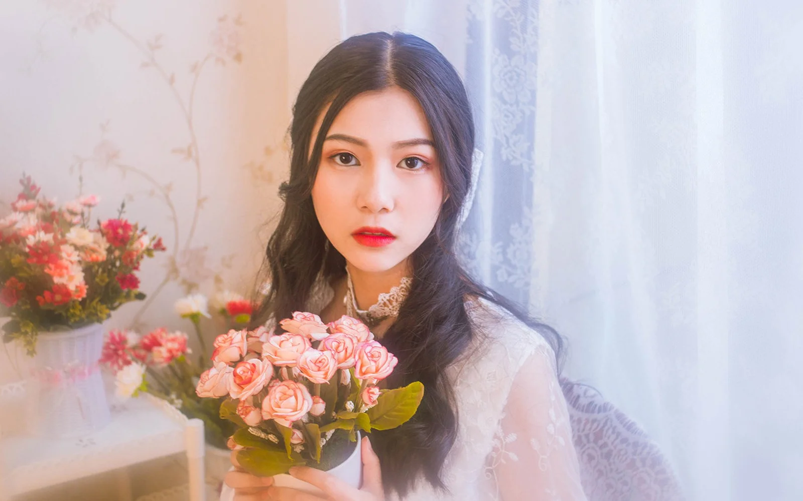 一名成年女性在室内手捧粉色花束，身后可见花卉与浅色窗帘。