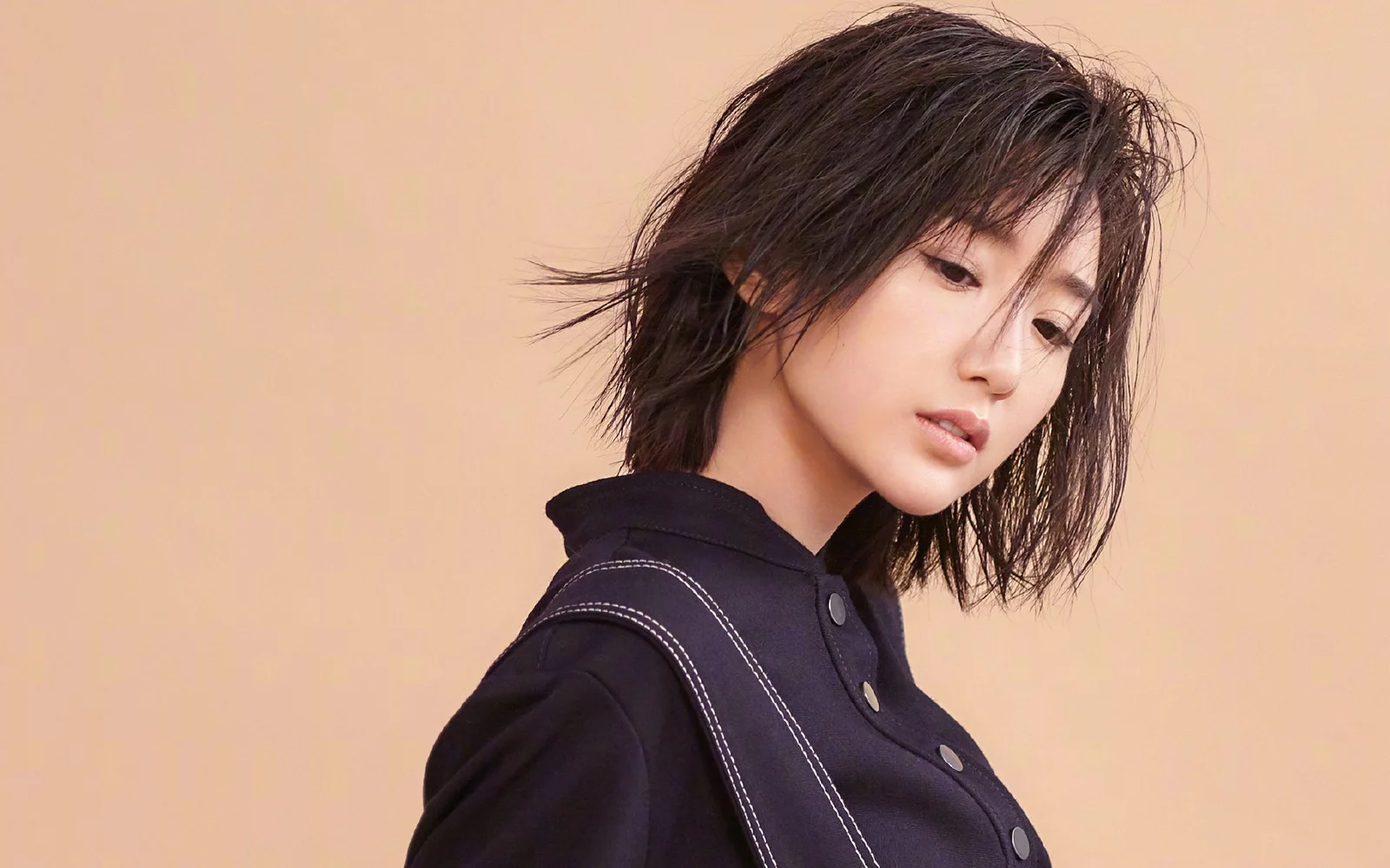 黒髪の女性が暗い服を着て、薄い茶色の背景の前でカメラの外を見つめています。