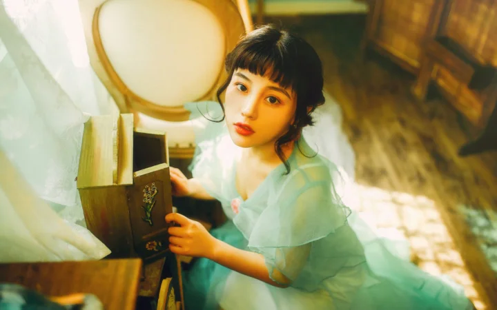 一名短发女性穿着浅绿色薄纱服装坐在木质家具旁，室内光线从侧面照入。