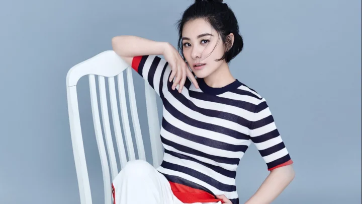 一名成年女性身穿黑白条纹上衣，倚靠在白色椅子旁的灰蓝色背景前。