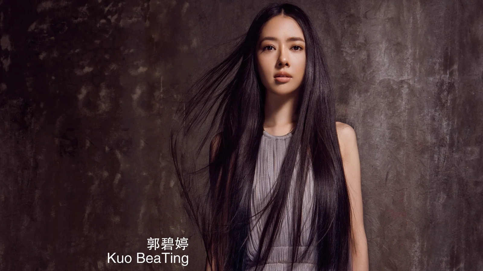 一名长发女性身穿灰色无袖上衣站在深色纹理背景前。
