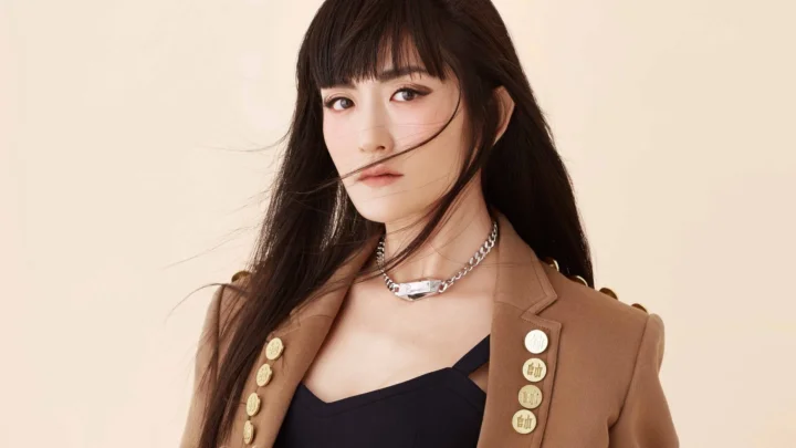 一位成年女性身穿棕色西装外套站在浅色背景前，呈现时尚人像画面。