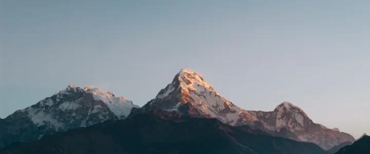 群山在柔和光线下延伸，主峰覆盖着积雪，背景是开阔的淡蓝色天空。