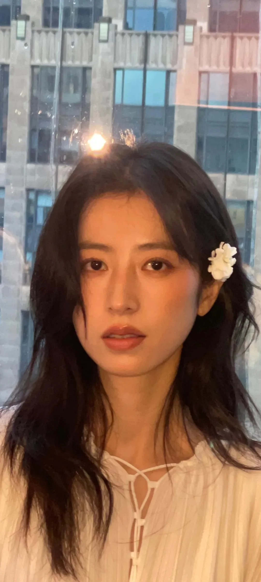 긴 검은 머리의 여성이 창가에서 카메라를 바라보고 있으며 뒤로 도시 건물이 보입니다.