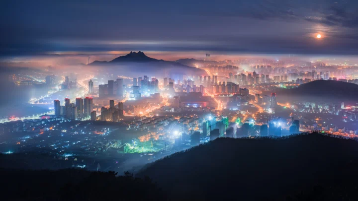 夜幕下灯火璀璨的城市天际线映衬着远处朦胧山峦，云层间可见明月。
