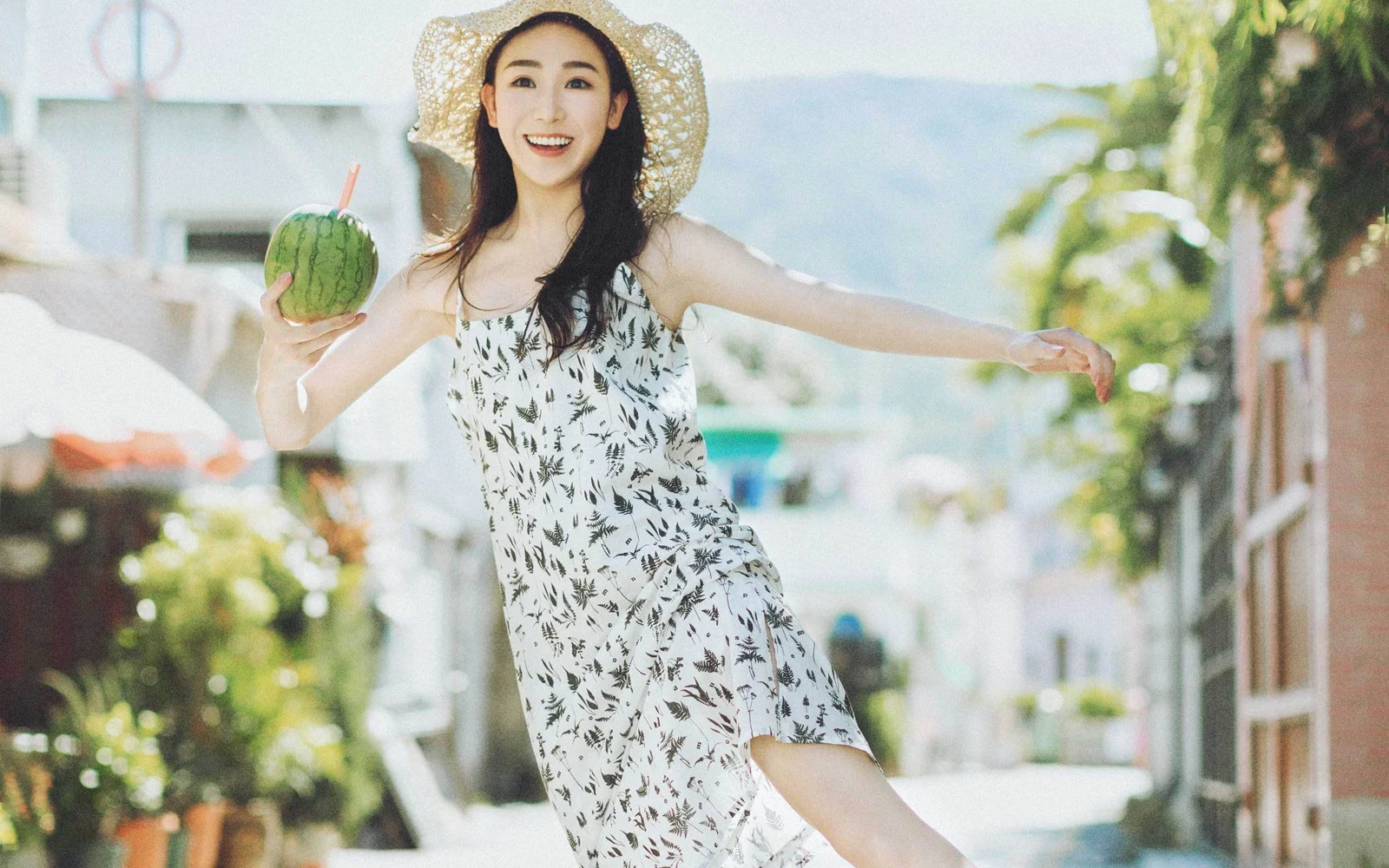 一位戴着草帽的女士穿着印花连衣裙站在阳光明亮的街道上，手里拿着一个绿色水果。