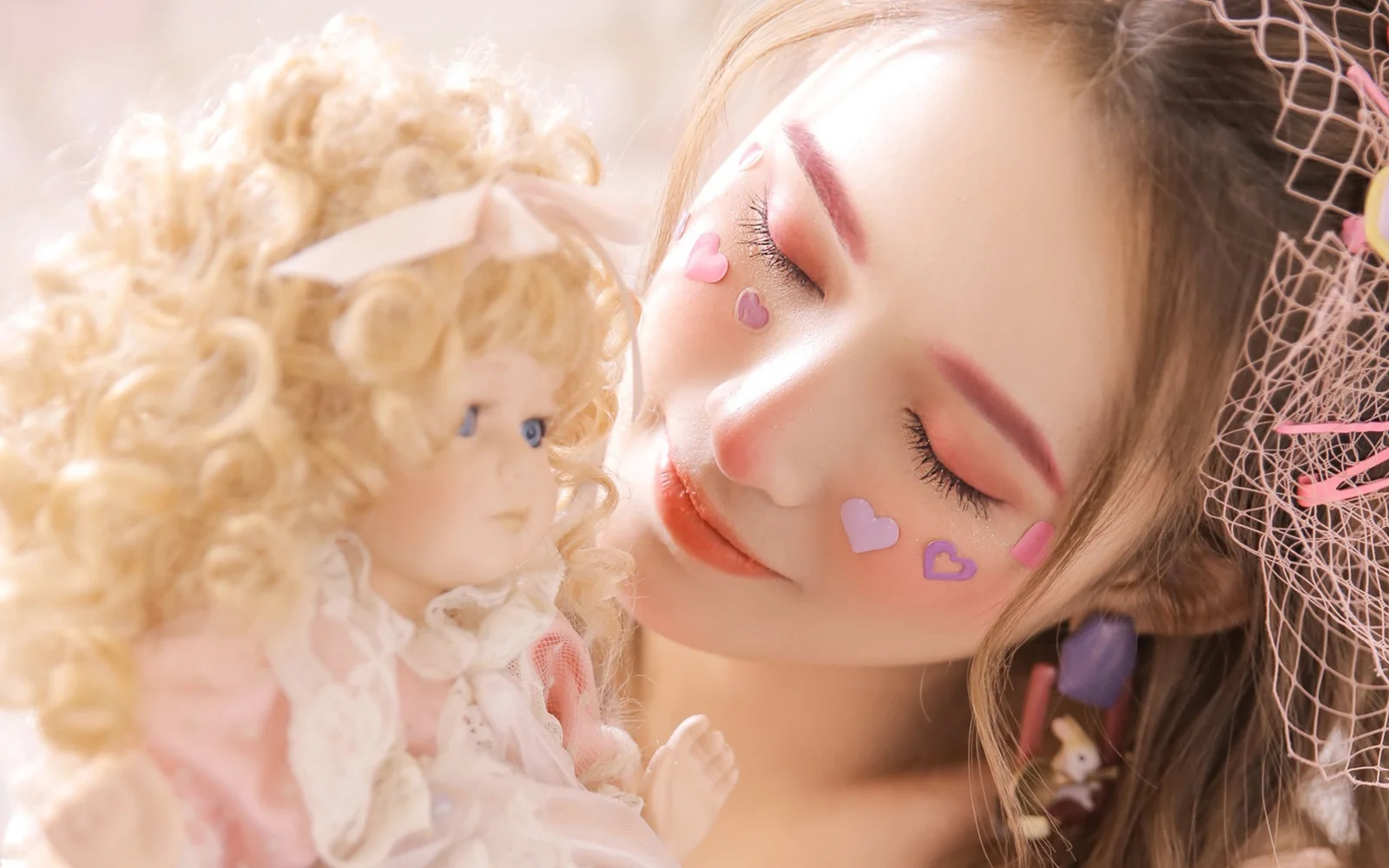 目を閉じた女性が金髪の人形に寄り添い、頬にピンクのハートシールを貼っている。