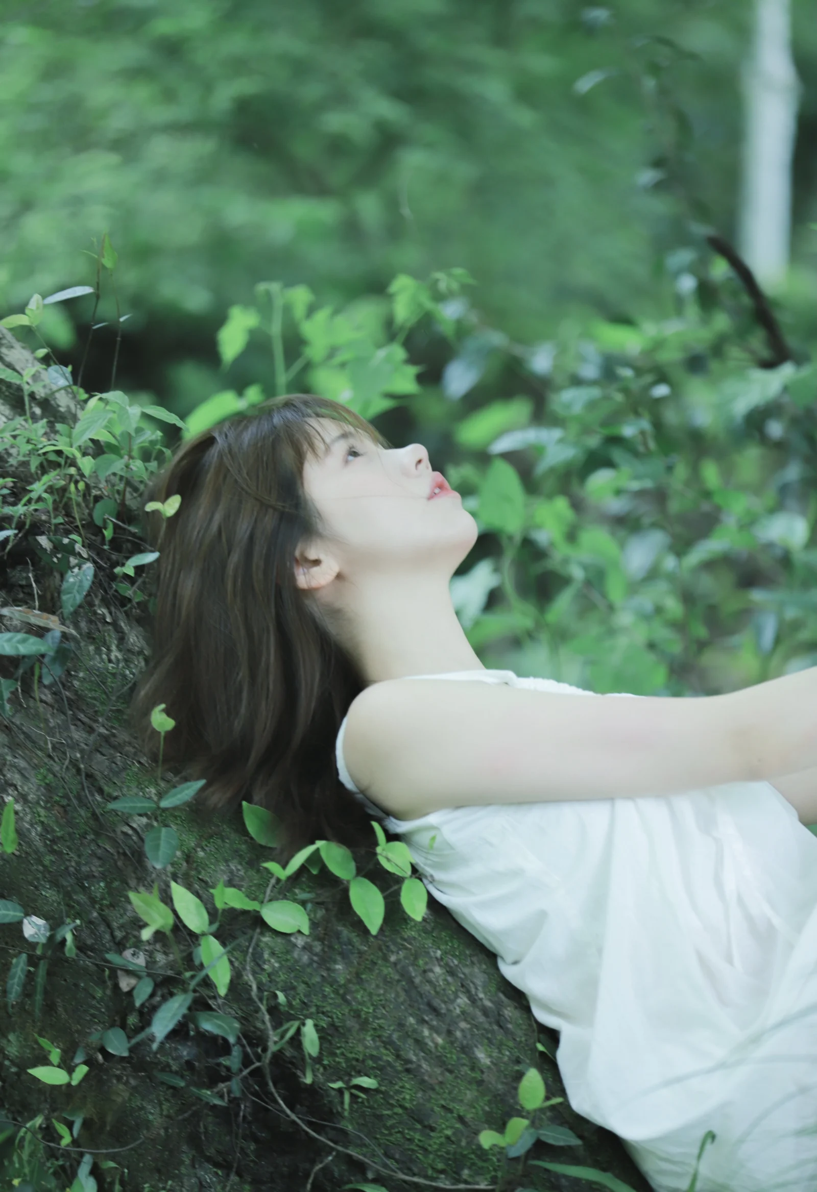 흰 드레스를 입은 여성이 푸른 숲의 식물 사이에 기대어 위를 바라보고 있습니다.