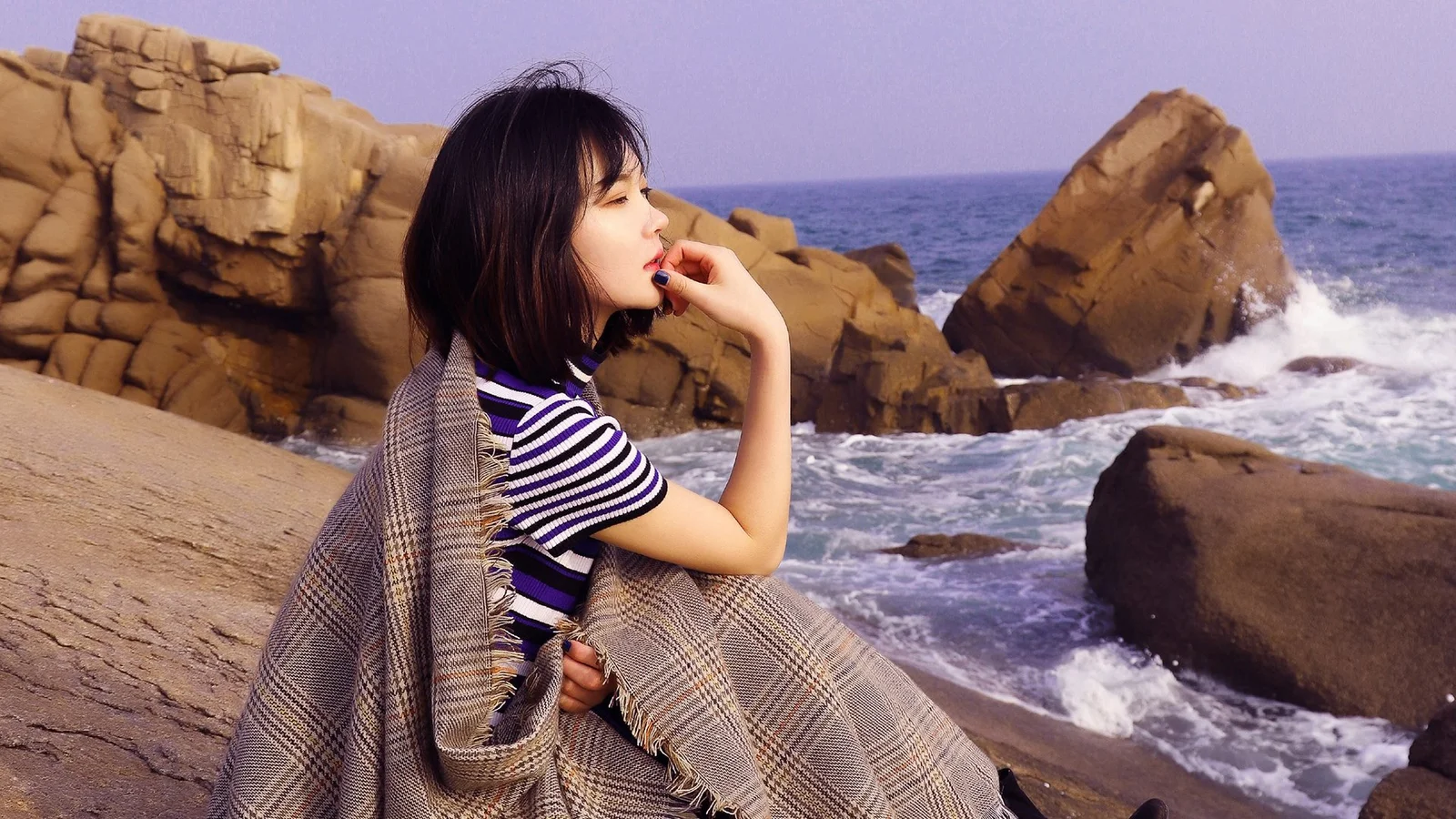 한 여성이 바닷가 바위에 앉아 있고 뒤로 파도와 바위 해안이 보인다.