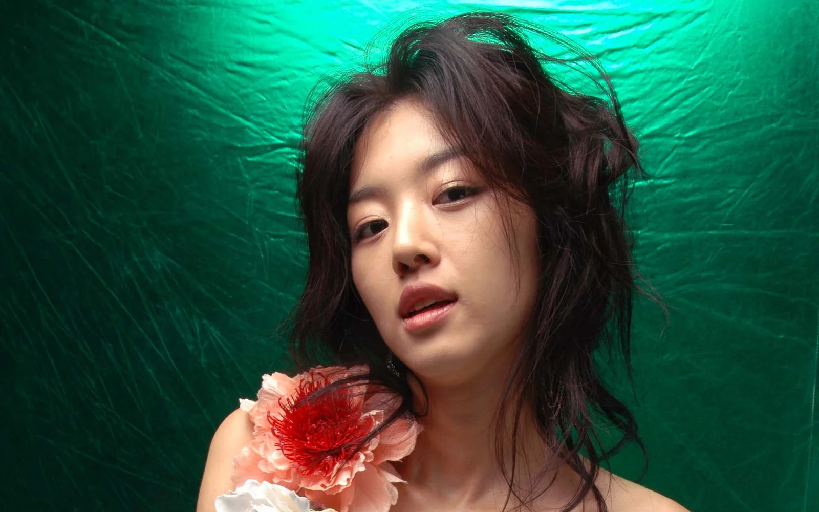 초록색 배경 앞에서 어깨 곁에 붉은색과 분홍색 꽃을 장식한 성인 여성이 카메라를 바라보고 있다.