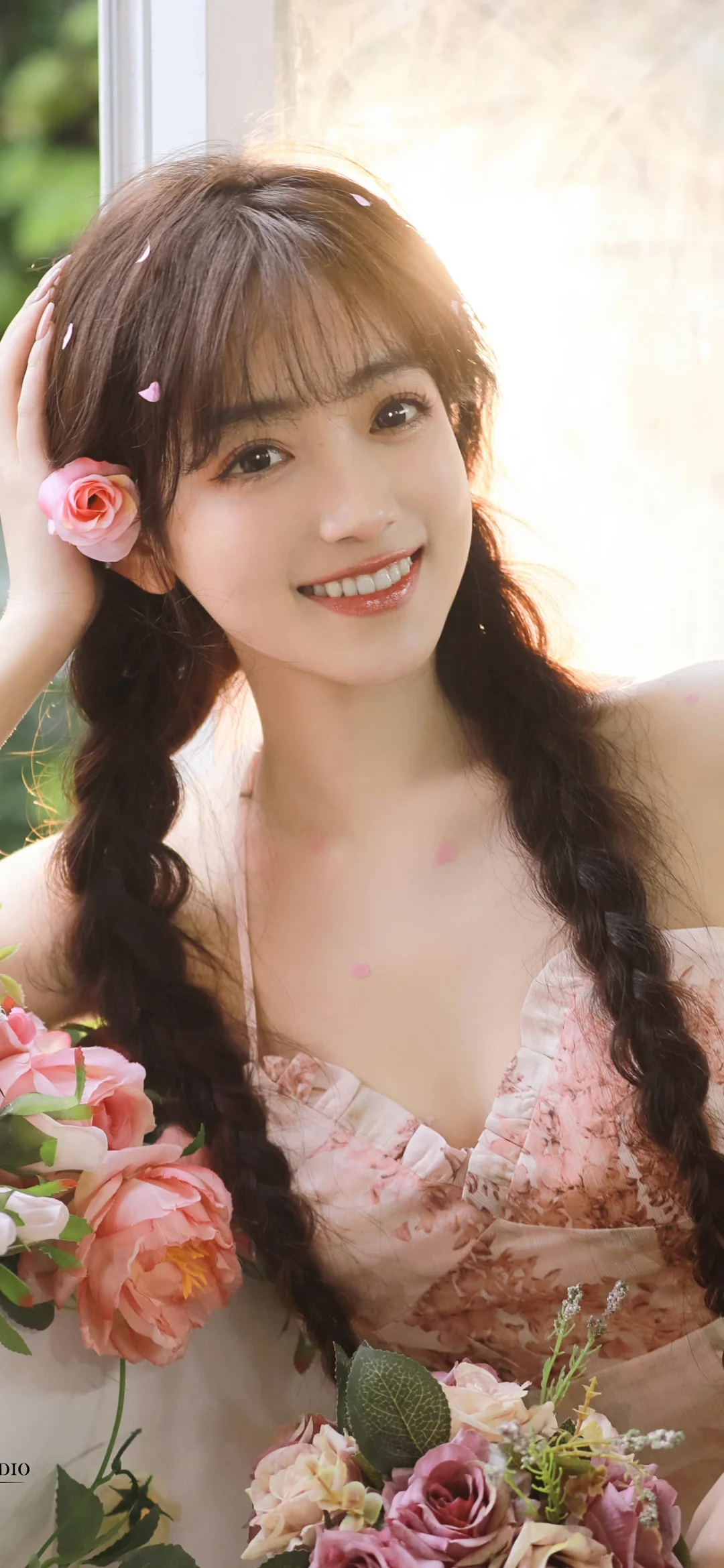 一名长发女子手持粉色花束，在明亮窗边微笑。