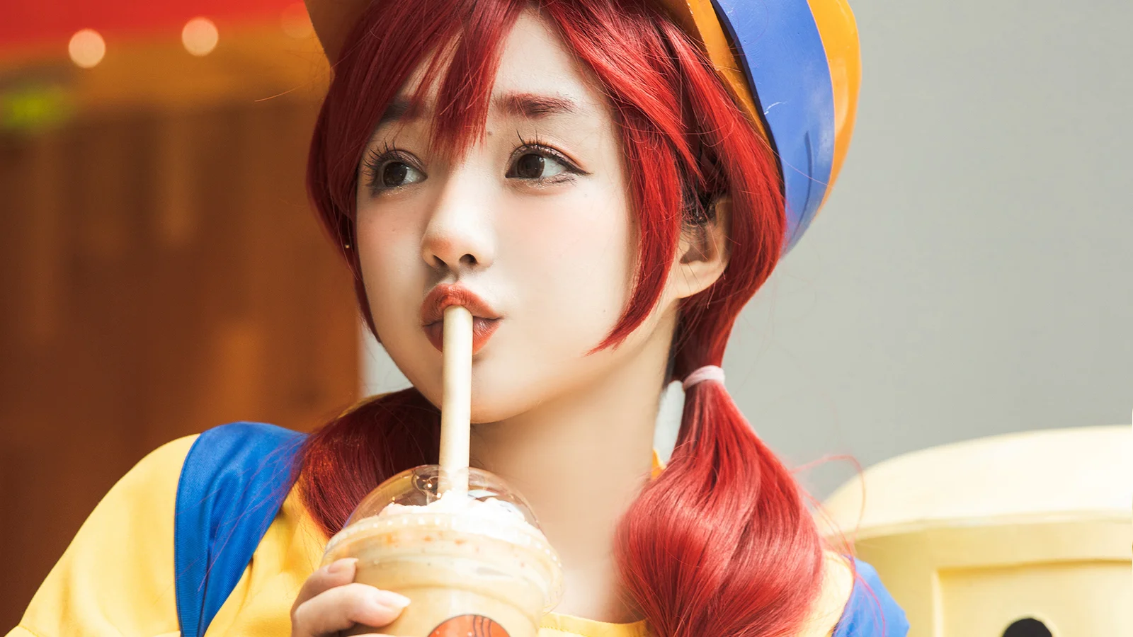 빨간 머리의 코스프레 인물이 밀크셰이크를 들고 옆을 바라보고 있다.