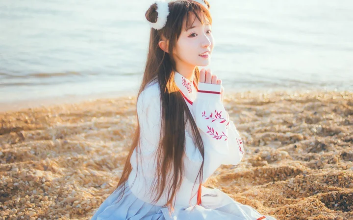 一名长发女子坐在阳光下的海边，身后是海水与碎石滩。