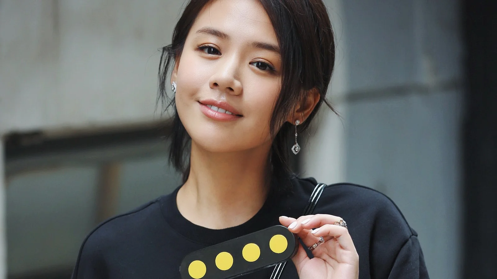 검은색 상의를 입은 성인 여성이 실내 배경 앞에서 미소를 지으며 노란색 원이 있는 검은 물체를 들고 있다.