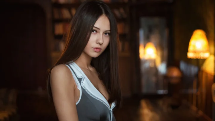 一名棕色长发女性身穿灰色无袖上衣站在昏暗温暖的室内灯光前。