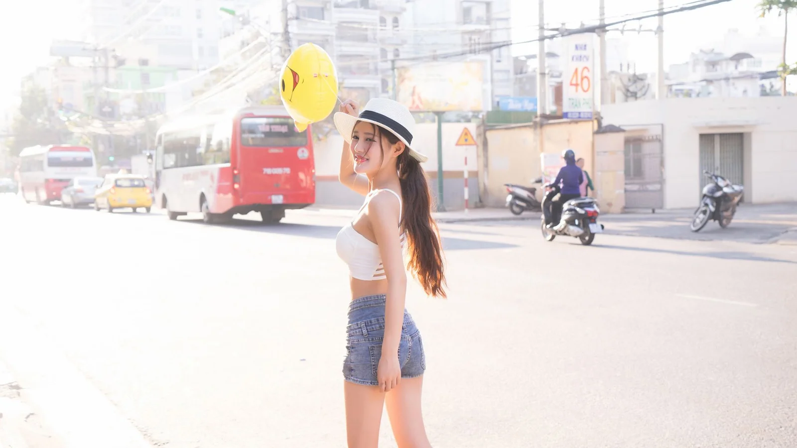 一名戴着帽子的年轻女子手持黄色气球站在阳光明亮的城市街道上。