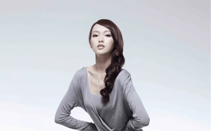 一名身穿灰色上衣、留着侧分长卷发的成年女性在浅灰背景前进行棚拍肖像。