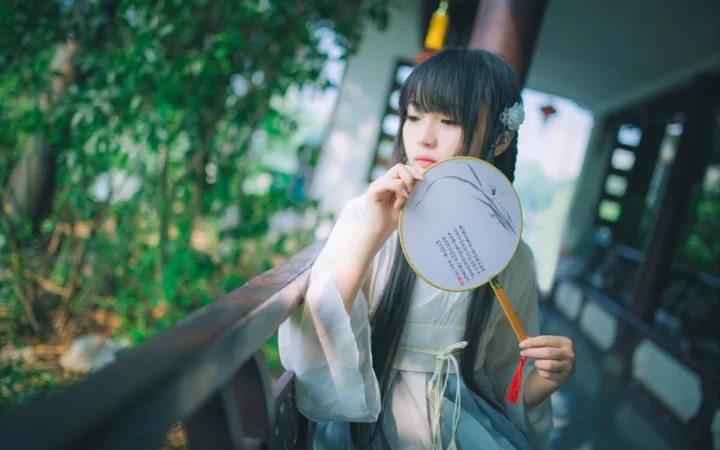 一名身穿浅色传统服饰的女子站在木栏旁，手持团扇望向镜头。