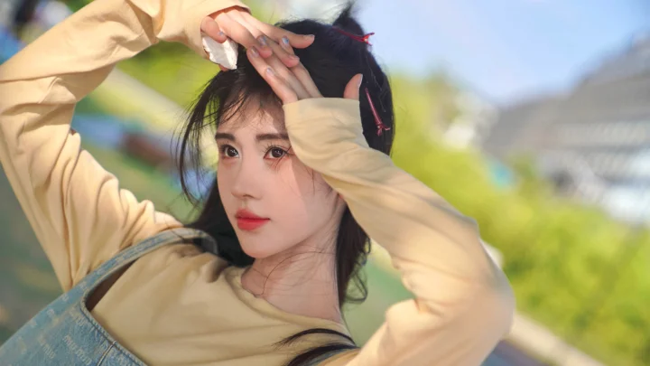一名黑发女子在户外阳光下抬手遮光，背景呈现柔和的绿色与建筑虚影。