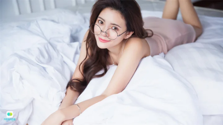 一名戴圆框眼镜的成年女子趴在白色床上看向镜头。