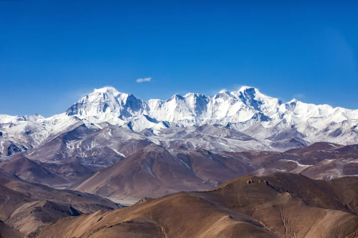 蓝天下，一片白雪覆盖的高山山脉横跨画面，前景是棕色山坡。