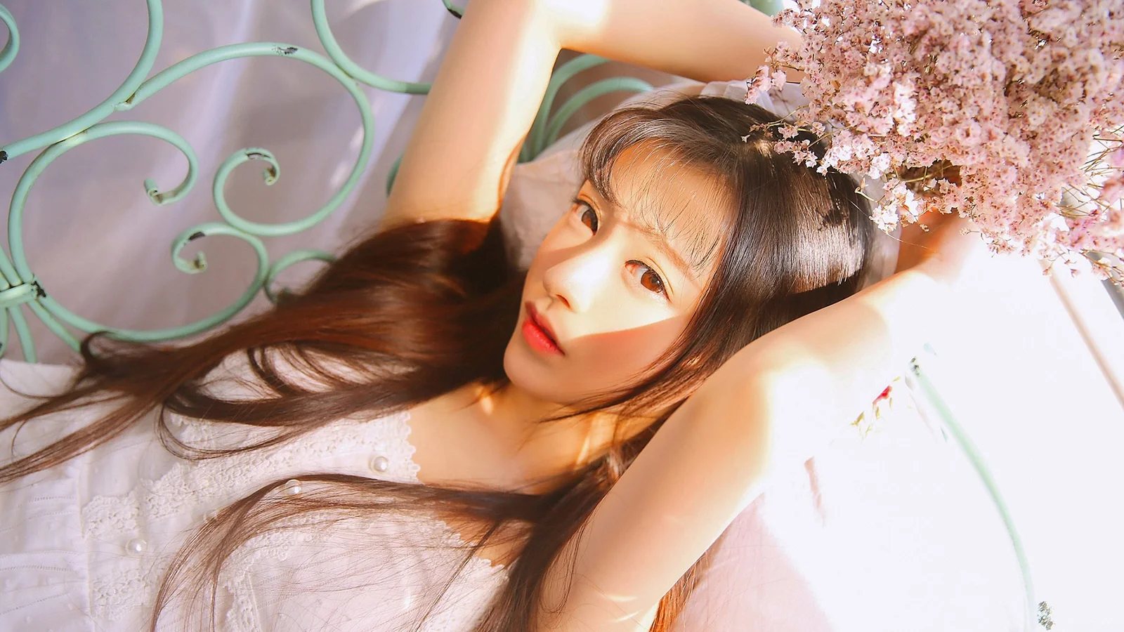 긴 머리의 여성이 부드러운 햇살 아래 누워 있으며 곁에 분홍색 꽃과 연한 초록색 장식이 보인다.