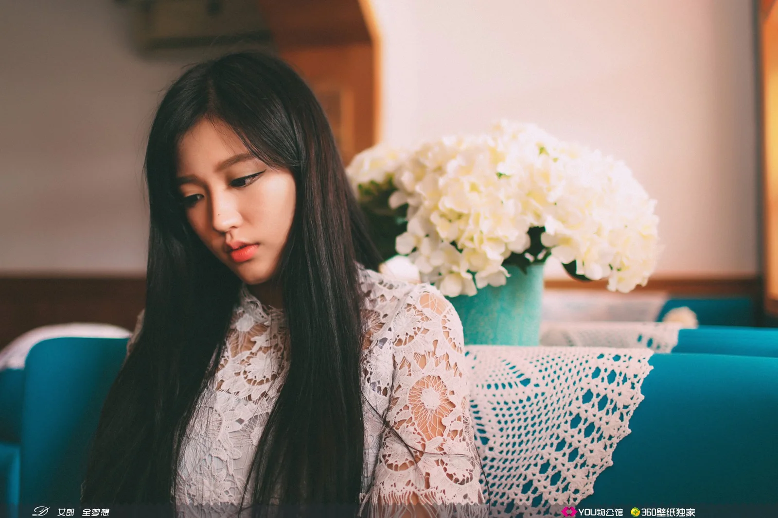 一位长发女性坐在室内沙发前，身后摆放着一盆白色花朵。