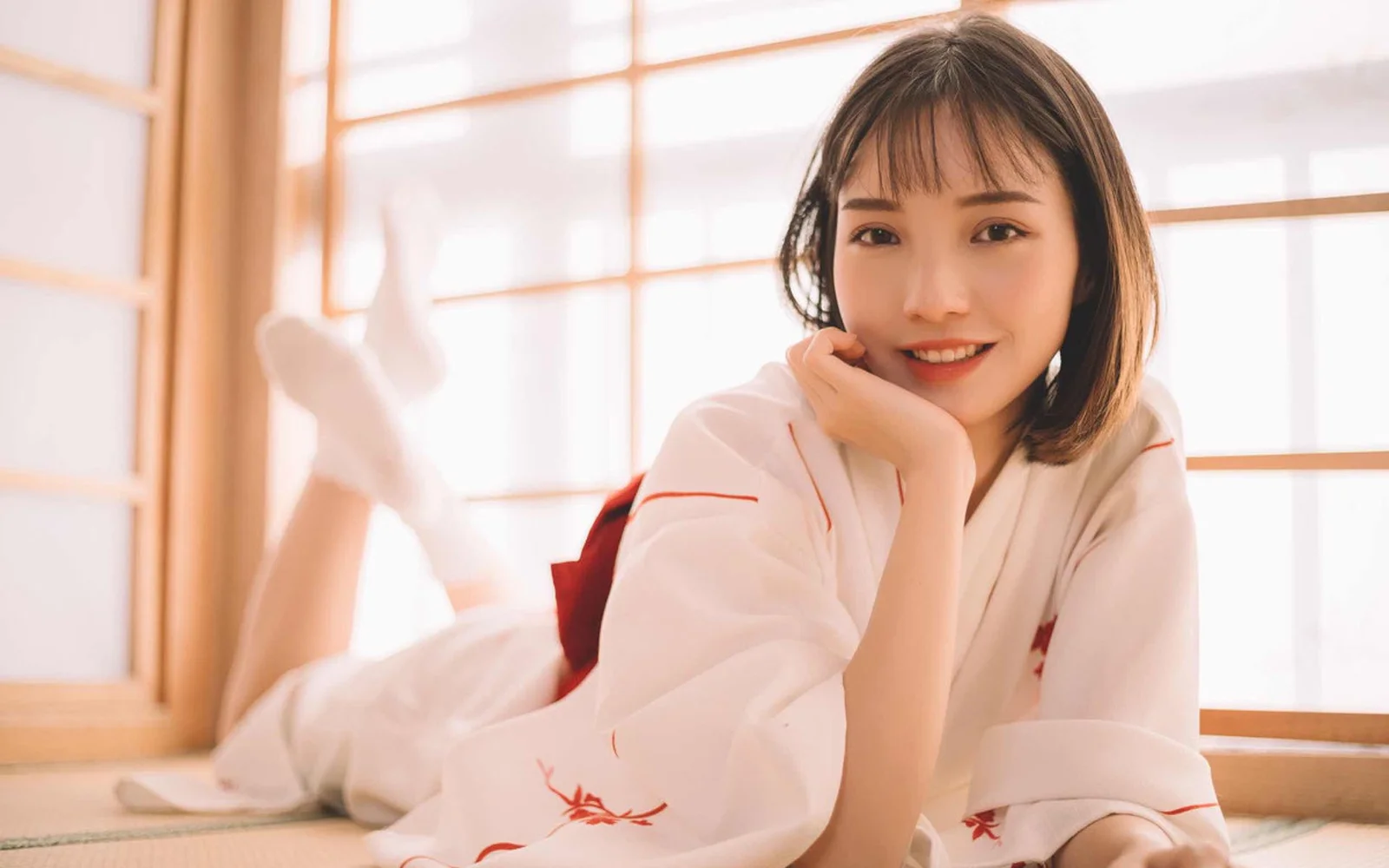 一名短发女性穿着白色衣服，趴在明亮窗边微笑看向镜头。