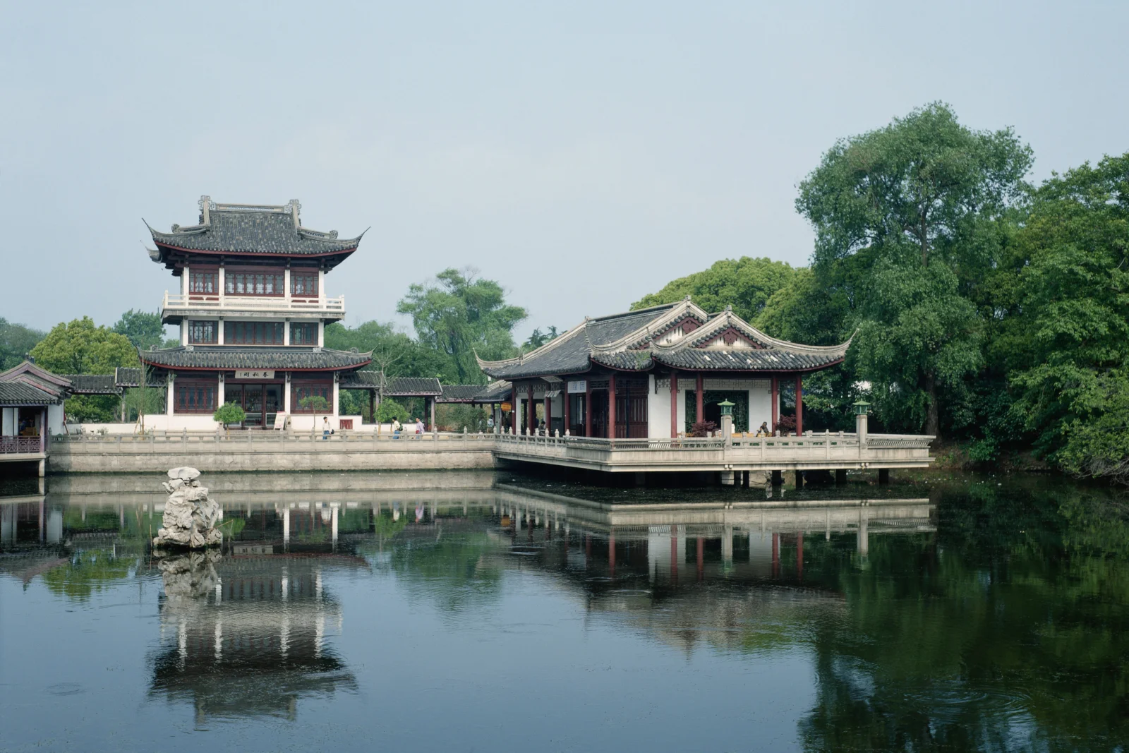 古典中式園林建築坐落在池塘邊，水面倒映著亭台、樹木與晴朗天空。