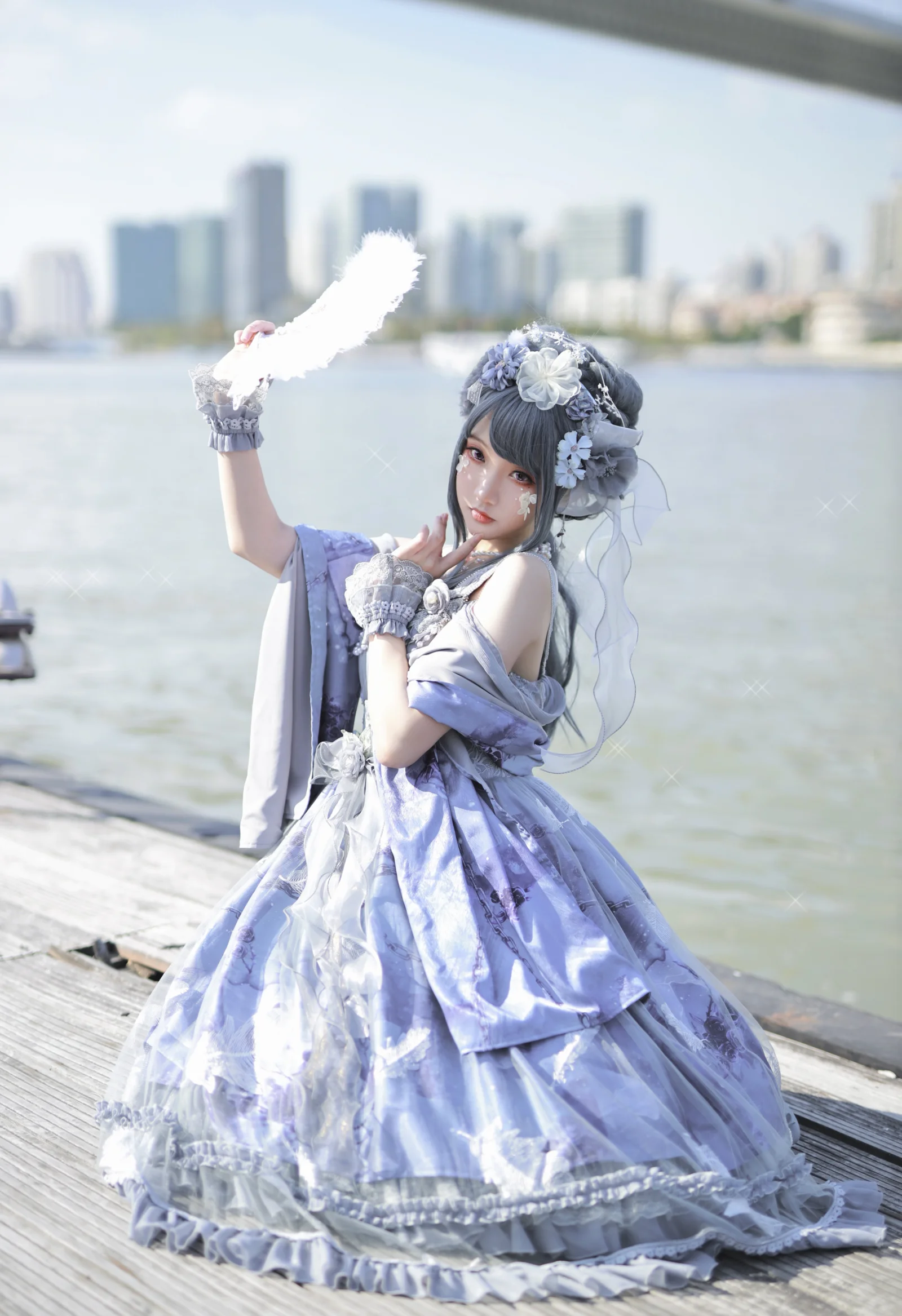 紫色の華やかなドレスを着た成人女性が、水辺の木道で白い羽根を掲げ、遠くに街のスカイラインが見えます。
