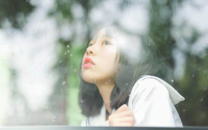 물방울이 맺힌 유리 너머로 한 여성이 흐릿한 초록빛 배경을 바라보고 있습니다.