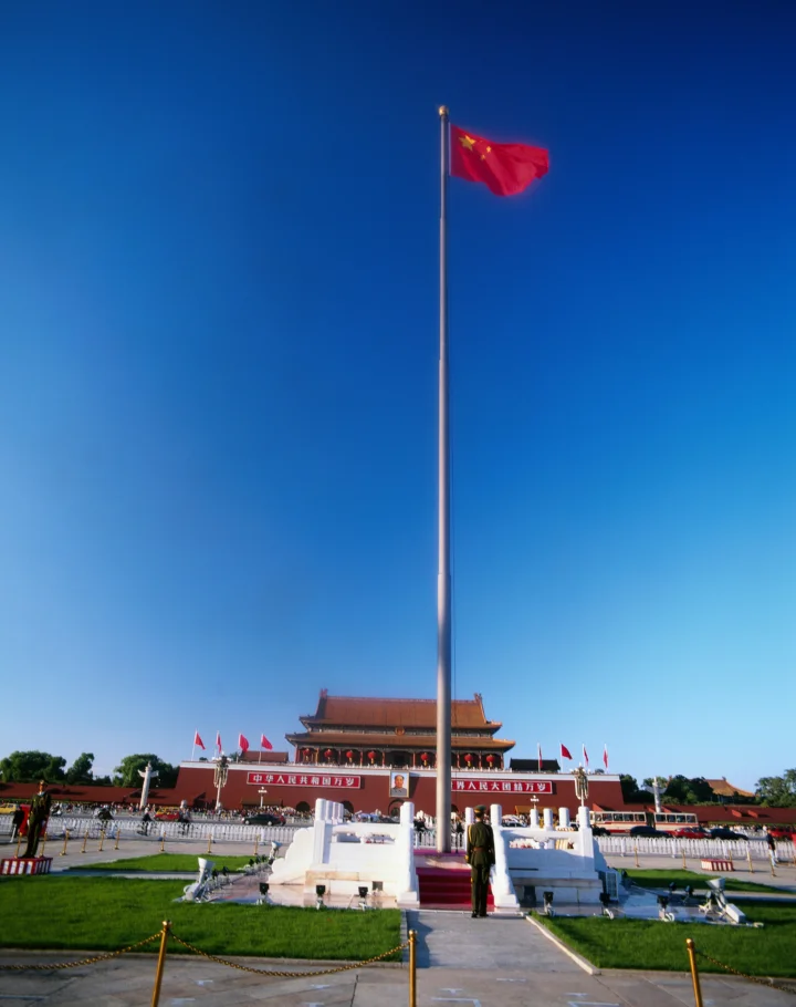 一面红旗在蓝天下飘扬，前方是宏伟的传统建筑和开阔广场。