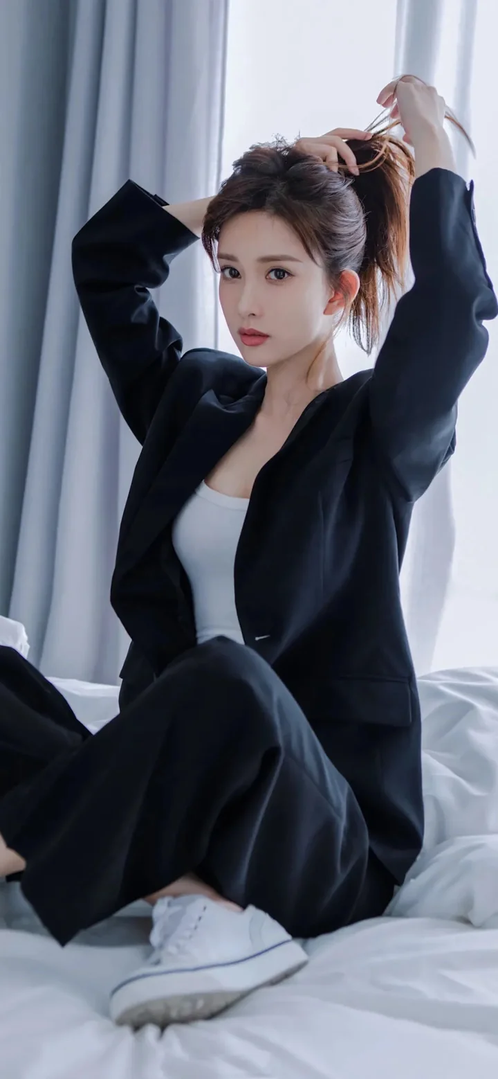一名成年女性身穿黑色西装坐在床上整理头发。